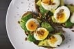 Close-up of avocado toast topped with boiled eggs, pine nuts, and seasoning on a white plate.