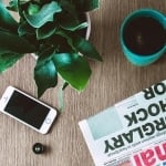 Top view of a wooden table with a green plant, a white smartphone, a teal coffee mug, and a folded newspaper